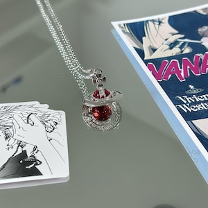 May include: A silver-toned necklace featuring a pendant shaped like a Saturn-style orb. The orb has a deep red spherical center surrounded by a metallic ring encrusted with small clear crystals and a small cross topper with a central crystal.
