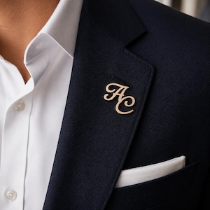 May include: A rose gold-toned metal lapel pin featuring the interconnected cursive letters AC, attached to the lapel of a dark navy blue textured blazer worn over a crisp white collared shirt with a white pocket square visible in the breast pocket.