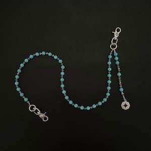 May include: A wallet chain featuring alternating turquoise-colored ribbed beads and small metallic spacers. The chain includes two silver-toned lobster claw clasps at the ends and a small octagonal charm with a compass rose design.