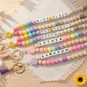 May include: A collection of five personalised lanyards featuring round silicone beads in pastel colours, white letter beads spelling names like MISS OLSEN, MRS HARVIE, BROWN, ESTHER, and SAMASON, yellow smiley face beads, and gold-toned metal clasps.
