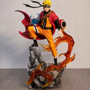 May include: A dynamic anime character figurine featuring a spiky blonde-haired figure in an orange and black outfit with a red cape. The figure stands on a gray rocky base surrounded by translucent orange flame effects and holds a blue energy sphere.