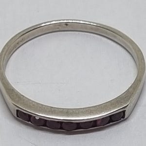 Puede incluir: Un anillo de metal plateado con un engaste de canal que presenta una fila de pequeñas piedras preciosas redondas de color rojo oscuro incrustadas a lo largo de la parte frontal de la banda. El anillo tiene un acabado liso y pulido.