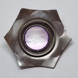 May include: A hexagonal silver-toned metal brooch featuring a central circular inlay of iridescent purple mother-of-pearl. The metal surface displays a series of etched semi-circular patterns radiating from the center toward each of the six corners.