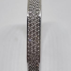 Könnte beinhalten: Ein silberfarbener Armreif mit einem breiten, flachen Band, das mit drei parallelen Reihen kleiner, klarer, rund geschliffener Steine besetzt ist. Das Armband enthält an der Basis einen runden Metallanhänger mit einem eingravierten Schwanenlogo.
