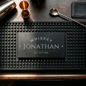 May include: A rectangular black rubber bar mat with a textured nub surface and a central smooth panel featuring the engraved text WHISKEY JONATHAN EST. MAY 1999. The mat is surrounded by a silver cocktail shaker, a copper jigger, and two long silver bar spoons.