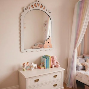 Peut inclure: Un miroir mural avec un cadre festonné blanc, doté d'un sommet en forme de couronne et d'une silhouette de château couleur cuivre dans le coin inférieur droit. Le miroir reflète la pièce environnante, y compris une commode et des objets décoratifs.