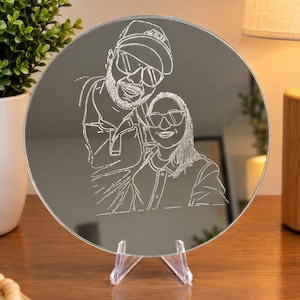 Peut inclure: Un miroir circulaire présentant une gravure au trait blanc représentant deux personnes portant des lunettes de soleil et des chapeaux. Le miroir est soutenu par un support en plastique transparent, affichant un portrait détaillé.