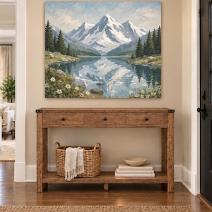 May include: A rectangular wooden console table with three drawers and a lower shelf. On the shelf sits a woven basket containing a white fringed cloth, a small bowl, and a stack of books. Above the table hangs a landscape painting of snow-capped mountains, a lake, and pine trees.