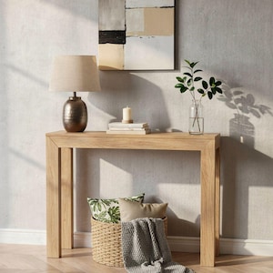 May include: A light oak console table featuring a dark metallic lamp with a beige shade, a stack of books, a white candle, and a glass vase with green foliage. A woven basket sits underneath, containing a patterned pillow and a gray knit throw blanket.