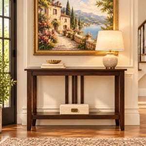 May include: A dark brown wooden console table with a lower shelf, featuring a rectangular top and four straight legs. On the table sits a beige lamp with a fabric shade and a stack of books with a gold bowl. A rectangular box sits on the shelf.