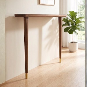 May include: A narrow console table constructed from dark brown wood with a smooth, rectangular top. The table features two long, tapered legs that extend to the floor, each finished with a polished gold-toned metal cap at the base.