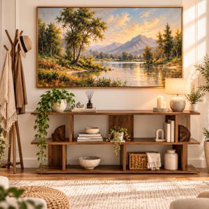 May include: A rectangular landscape painting in a gold frame depicting a mountain lake at sunset, hung above a long wooden console table with three shelves. The table holds a potted plant, books, a woven basket, and a table lamp.