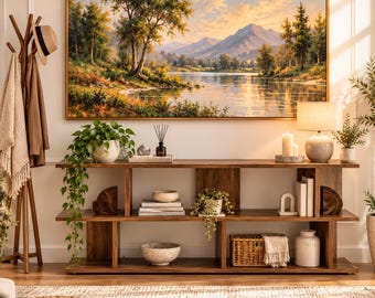 May include: A rectangular landscape painting in a gold frame depicting a mountain lake at sunset, hung above a long wooden console table with three shelves. The table holds a potted plant, books, a woven basket, and a table lamp.