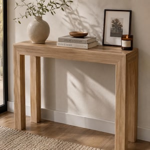 May include: A light oak wood console table with a rectangular top and four straight legs. On the table sits a textured beige vase with green branches, two stacked books titled Live Beautifully and This Is Home, a small wooden bowl, a framed print, and a candle.