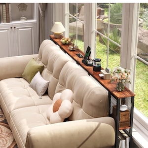 May include: A long, narrow wooden console table with a black metal frame positioned behind a beige tufted sofa. The table surface holds a wireless charger, a small lamp, a decorative vase with flowers, and a tissue box, with storage shelves below.