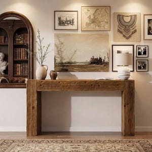 May include: A rectangular wooden console table with a natural grain finish, featuring a thick top and sturdy block legs. The table is positioned against a wall decorated with various framed art prints, a map, and a white table lamp with a wavy base.