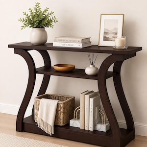 May include: A dark brown wooden console table with curved side supports and a middle shelf. The top surface holds a potted plant, two stacked books labeled "LIVE BEAUTIFULLY" and "THIS IS HOME", a picture frame, and a candle. The lower shelf holds a woven basket with a white cloth, and three books held by two marble bookends.