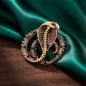 May include: A gold-toned metal brooch shaped like a coiled cobra snake. The snake features a textured scale pattern and is accented with small, round, vibrant green rhinestones embedded along its body and in the eye of the cobra head.
