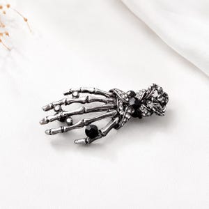 Może przedstawiać: Broszka z metalu w kolorze srebrnym w kształcie szkieletowej dłoni. Element posiada teksturowane wykończenie przypominające kość, ozdobione małymi przezroczystymi cyrkoniami wzdłuż palców i nadgarstka oraz kilkoma większymi, okrągłymi, fasetowanymi czarnymi kamieniami.