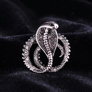 May include: A silver-toned metal brooch shaped like a coiled cobra snake. The snake features a raised hood, textured scales, and small clear crystals embedded along the circular body. The cobra has a dark, polished finish with black eye details.