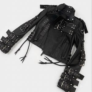 May include: A black leather jacket featuring multiple silver-toned metal buckles and straps along the sleeves and chest. The garment includes side lacing with dangling cords, a notched collar, zippered pockets, and metallic stud embellishments.