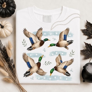 May include: A white crewneck sweatshirt featuring a graphic print of four mallard ducks in flight. The ducks have brown, white, and iridescent blue wing feathers with green heads. The design includes light blue horizontal brush strokes and small leaves.