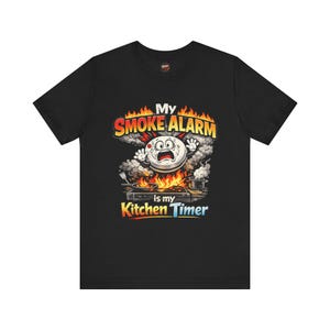 May include: A black short-sleeved t-shirt featuring a graphic of a panicked smoke alarm character above a stove with flames and smoke. The text reads: My SMOKE ALARM is my Kitchen Timer. The design includes orange, yellow, and grey colours.