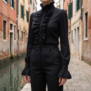 Peut inclure: Une chemise noire à manches longues dotée d'un col montant à volants, d'une patte de boutonnage verticale à volants sur le devant et de poignets évasés à double épaisseur de volants. La chemise est portée avec un pantalon gris foncé.