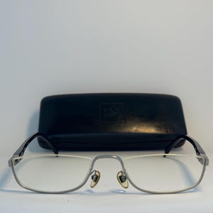 May include: Rectangular eyeglasses with a silver metal frame and black temples, featuring clear lenses and gold-toned nose pads. A black hard-shell glasses case with the embossed text "alain mikli" is positioned directly behind the eyewear.