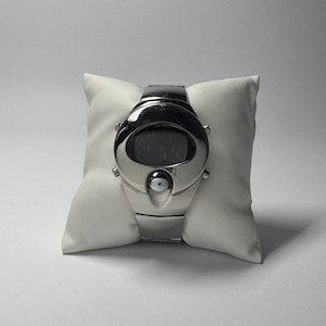 May include: A silver-toned digital wristwatch featuring a smooth, rounded, futuristic case design with a dark display screen and a circular button detail below the face. The watch is secured with a matching metallic link band and rests on a white pillow.