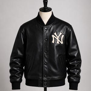 Pode incluir: Um casaco varsity de couro preto com gola, punhos e bainha canelados em preto. A parte frontal apresenta um fecho de botões de pressão e um proeminente logótipo dos New York Yankees bordado a branco e preto no lado esquerdo do peito.