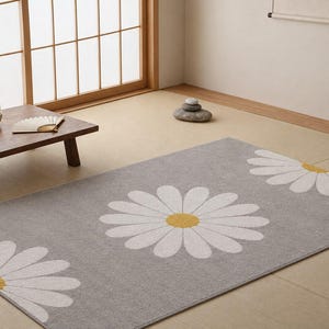 May include: A rectangular area rug featuring a light gray background with a repeating pattern of large white daisy flowers with yellow centers. The rug has a soft, textured surface and clean, finished edges, providing a neutral floral design.