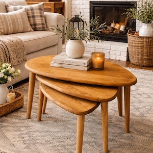May include: A set of three nesting tables made of light brown wood with smooth, rounded, teardrop-shaped tops and tapered legs. The tables are stacked together, with a white ceramic vase, two books, and a glass candle holder placed on top.
