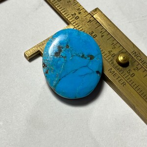 Peut inclure: Une pierre précieuse turquoise de forme ovale avec une couleur bleu vif et des veines de matrice brun foncé. La pierre est placée à côté d'un pied à coulisse en laiton, montrant qu'elle mesure environ 3,2 cm de longueur et de largeur.