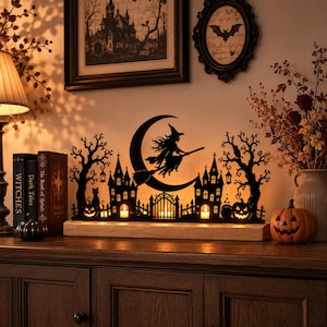 Op de afbeelding: Een zwarte metalen silhouetdecoratie met een heks op een bezemsteel vliegend voor een maansikkel, omringd door spookkasteeltorens, kale bomen en pompoenen, gemonteerd op een lichte houten voet met warme interne verlichting.