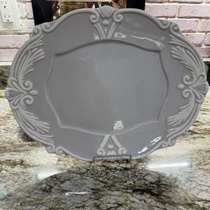 May include: A light gray oval ceramic serving platter featuring a smooth central surface and a raised, ornate relief border with symmetrical scrollwork and leaf-like patterns along the edges, supported by a small metal display stand.
