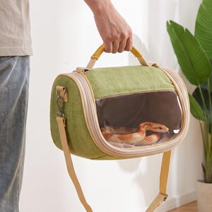 Peut inclure: Un sac de transport pour animal de compagnie vert avec une fenêtre de visualisation en plastique transparent sur le devant. Le sac dispose d'une poignée supérieure jaune et d'une bandoulière réglable jaune assortie. Un serpent orange et blanc est visible.