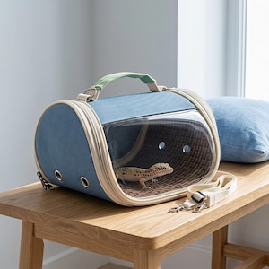 Puede incluir: Un transportín azul para mascotas con panel frontal de plástico transparente, asa superior verde claro y bordes color crema. Un pequeño gecko leopardo moteado es visible en el interior. Una correa de hombro color crema desmontable está al lado.
