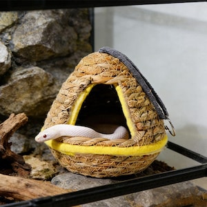 May include: A woven straw dome-shaped pet hideout with a triangular entrance trimmed in bright yellow fabric. A grey fabric strap with a metal ring is attached to the top. A white snake with red eyes is partially visible inside the structure.