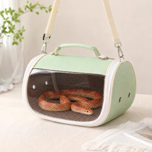 May include: A light green cylindrical pet carrier with a clear front window, a top handle, and an adjustable shoulder strap. Inside the carrier, a patterned orange and tan snake rests on a dark brown textured base, with metal ventilation grommets.