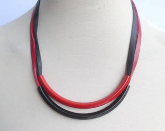 May include: A red and black leather necklace with a curved design. The necklace is displayed on a white mannequin.