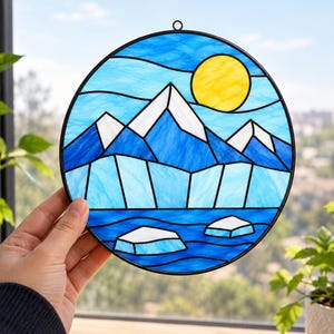 Op de afbeelding: Een ronde glas-in-lood zonnevanger met een landschap van blauwe bergen met witte toppen, een heldere gele zon en drijvende witte ijsbergen in een diepblauwe zee, omlijnd door een zwart metalen frame met een kleine ophangring aan de bovenkant.