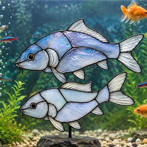 Könnte beinhalten: Zwei Buntglas-Fischskulpturen mit irisierenden blauen und weißen Paneelen, montiert auf einem dunklen Steinsockel. Die Fische sind übereinander positioniert, mit dunklen Umrandungen und klaren Flossen, ausgestellt in einem Aquarium.