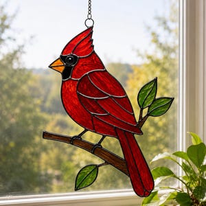 May include: A stained glass suncatcher shaped like a northern cardinal bird with vibrant red glass, a black face mask, and an orange beak. The bird is perched on a brown glass branch with three green glass leaves, suspended by a metal chain.