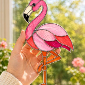 Op de afbeelding: Een glas-in-lood flamingo suncatcher met diverse tinten roze, rood en oranje glas, samengevoegd door zilverkleurige metalen soldeerlijnen. De flamingo heeft een wit met zwarte snavel en een klein zwart glazen oog en staat op één poot.