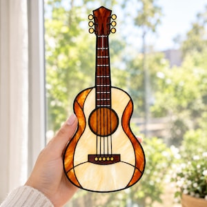 May include: A stained glass suncatcher shaped like an acoustic guitar, featuring cream-coloured panels for the body, dark amber edges, and a brown neck with black metal lead lines outlining the fretboard, sound hole, and bridge structure.
