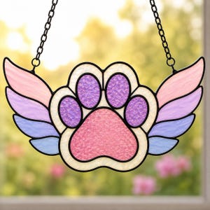 May include: A stained glass suncatcher shaped like a paw print with wings. The central paw pad is pink, surrounded by purple toe pads and white borders. The wings feature a gradient of pink, purple, and blue glass segments, all held by black metal.