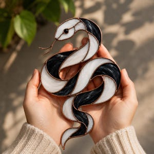 Pode incluir: Um ornamento de vitral em forma de serpente com segmentos alternados de vidro branco opaco e vidro preto marmorizado. As peças de vidro são unidas por finas linhas de solda metálica cor de cobre, formando uma serpente enrolada.