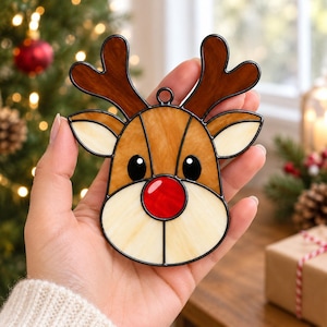 May include: A stained glass reindeer ornament featuring dark brown antlers, light brown ears, a cream-colored face, black eyes, and a bright red nose, all outlined in black metal solder with a small loop at the top for hanging.