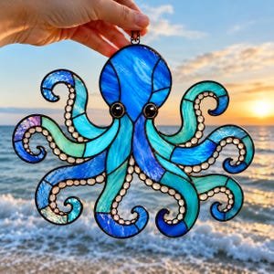 Könnte beinhalten: Ein Oktopus-Sonnenfänger aus Buntglas mit einer lebendigen Palette aus tiefblauen, türkisfarbenen und hellen blaugrünen Glassegmenten. Der Oktopus hat acht geschwungene Tentakel, die mit kleinen, klaren Glasperlen und zwei schwarzen Augen besetzt sind.